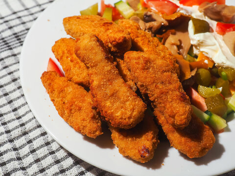 Golden, crispy breaded fried chicken pieces glow under warm light. They rest on a plain white plate and looking freshly cooked. Each piece has a textured, crunchy exterior. Fine goujons ready to eat