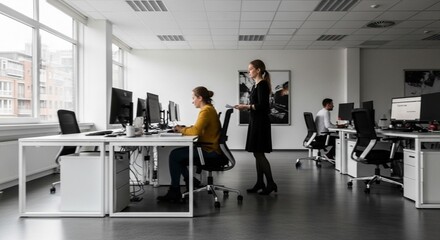 Fototapeta premium A modern office space with large windows, featuring three individuals engaged in work, with two women and one man, one woman standing and conversing with a seated colleague.
