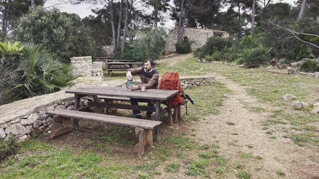 Man resting at picnic table drinking from mug during hiking trip.