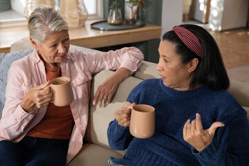 Diverse senior female friends sitting on beige sofa holding matching tan ceramic mugs and chatting © wavebreak3