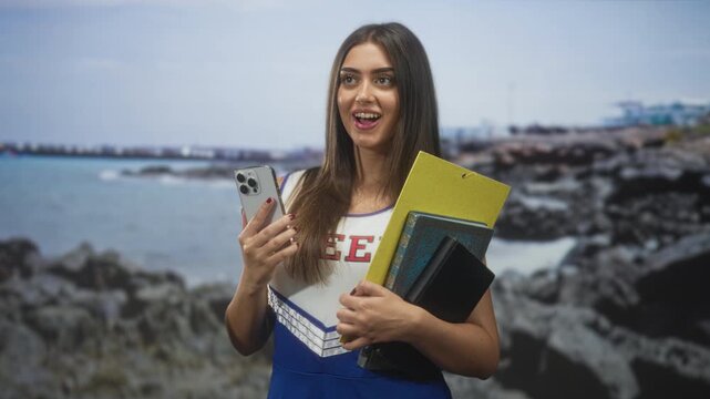 Woman holding smartphone and notebooks, smiling while looking up in a studio seafront set with rocky shore and pier visible; confidence ambition study.