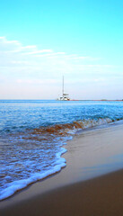 Fototapeta premium Sea waves at sandy beach and sailing catamaran moored at pier in background. Catamaran is used for cruises and charters to Asian resorts. Sailing catamaran anchored in calm waters at shore.