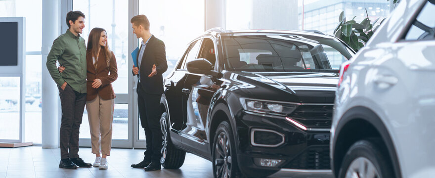 A couple is discussing their choice of a new car with a dealer in a modern dealership. The atmosphere is busy, with cars displayed to showcase various models.