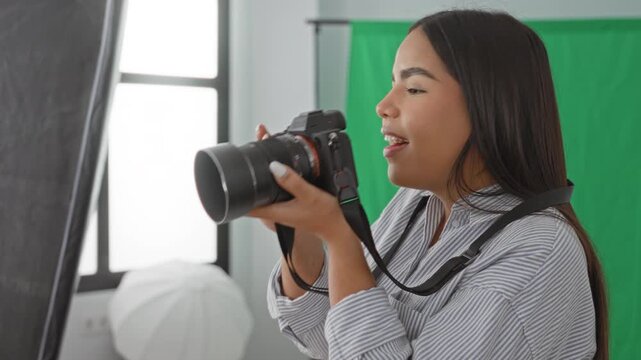 Woman holding camera with hands on lens, looking through viewfinder and later checking playback while smiling in studio with green screen and softbox; creative joy.