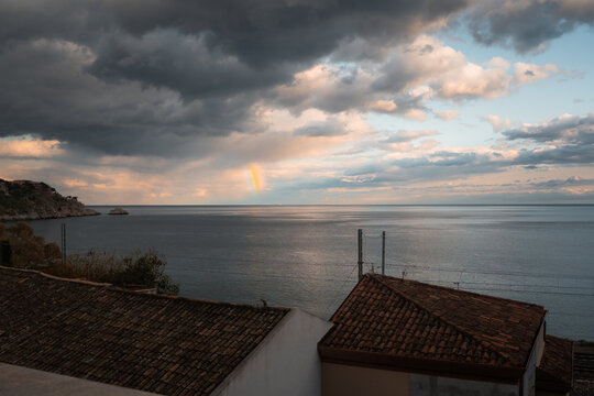 View of a faint rainbow arcing over the tranquil sea under a dramatic, cloud-streaked sky, terracotta rooftops adding warmth to the coastal scene, Taormina, Sicily, Italy.