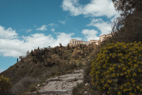 View of ancient stone stairs climbing toward a hilltop village amidst lush green trees against a backdrop of blue sky and white clouds, Taormina, Sicily, Italy.