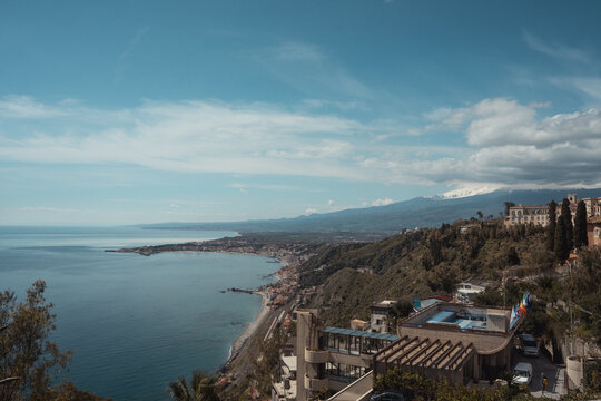 View of the azure sea meeting the rugged coastline under a vast sky, with buildings nestled among lush greenery, Taormina, Catania, Sicily, Italy.