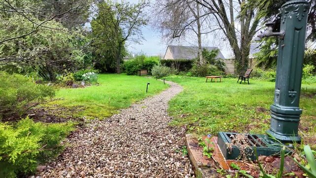 Low angle shot of a winding gravel path in a lush French garden featuring a vintage green water pump.