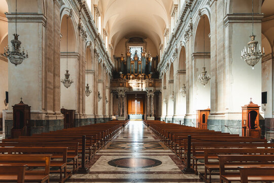 View of serene church interior with symmetric rows of wooden pews and ornate chandeliers casting shadows on the marble floor, Taormina, Sicily, Italy.