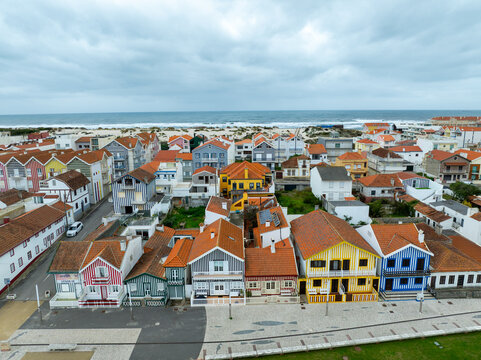 Aerial view of colorful houses with red and orange roofs meet sandy shores under a diffused sky, Costa Nova, Portugal.