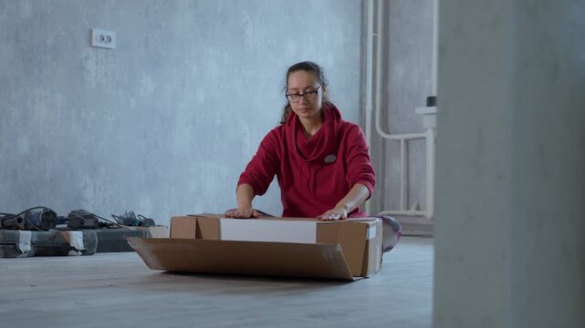 Female Organize Items During Renovation. Woman Arranges Cluttered Tools In Empty Room During Renovation. Asian Woman Carefully Packs Boxes In Barren Room Filled With Scattered Renovation Tools