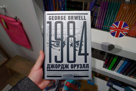 Minsk, Belarus. Apr 12, 2026. Iconic dystopian novel 1984 by George Orwell in both English and Russian languages held by customer in bookshop with many titles on white shelves, bilingual reading