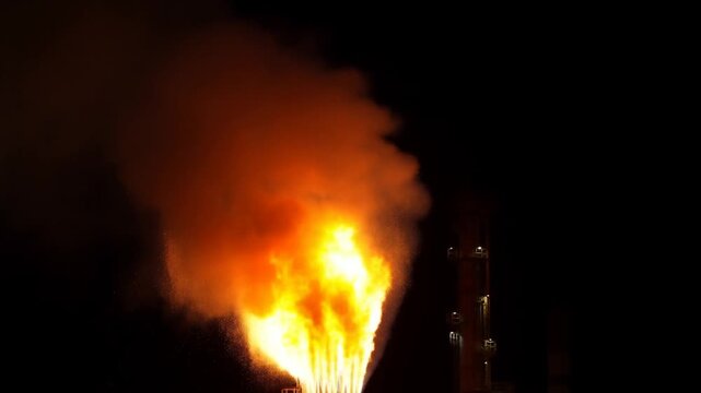 Static shot of industrial flare stack emitting large flames and thick smoke at oil refinery plant complex during night