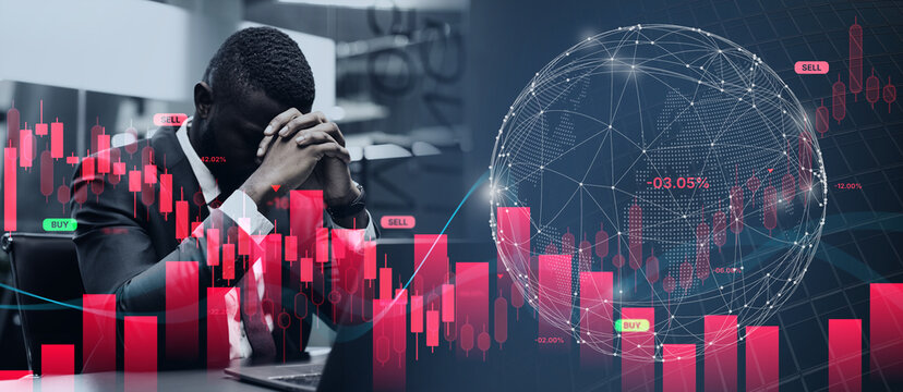 A businessman sits at his desk with his head in his hands, focused on a laptop displaying stock market charts. Red and green graphs show trading data in an office environment.