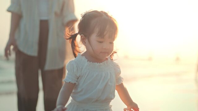 Happy Asian toddler girl running on the beach with her mother during sunset. Joyful child enjoying summer vacation and outdoor wellness activity by the sea, Beautiful golden hour natural light