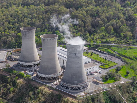 Larderello, Italy - 10 April 2026: Aerial view of Enel Green Power - Centrale Geotermica di Larderello with three large cooling towers emitting steam surrounded by lush green forests.