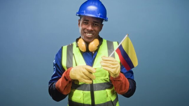 Man construction worker points finger to colombian flag while wearing hardhat and reflective vest in studio; pride solidarity.