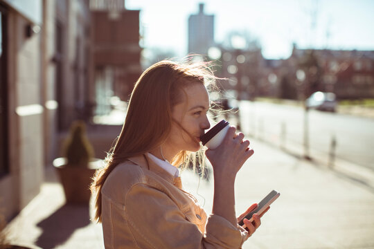Young woman drinking coffee and using smartphone on city street