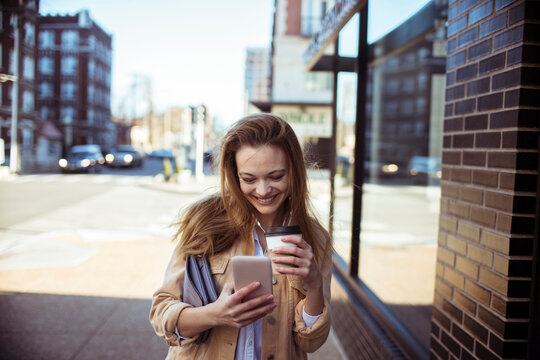 Smiling young woman using smartphone while holding coffee on city sidewalk