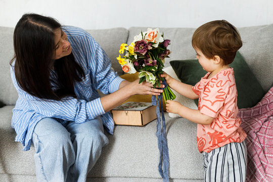 Cute child boy giving bouquet to beautiful mother on mothers day.  Happy Mother's day! Stylish Mom and son spending time together on couch at home, family holidays