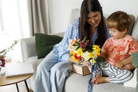 Cute child boy giving bouquet to beautiful mother on mothers day.  Happy Mother's day! Stylish Mom and son spending time together on couch at home, family holidays