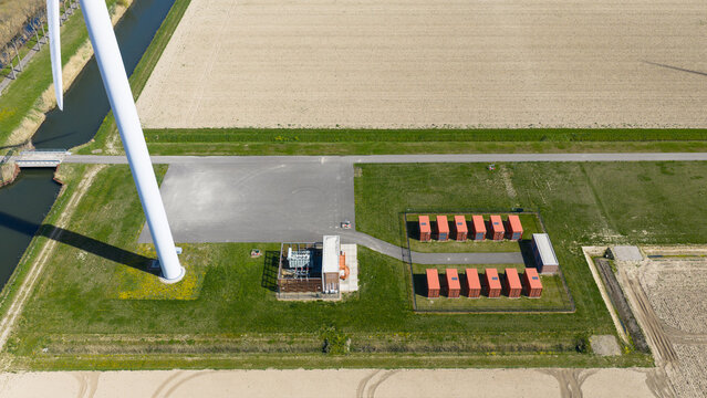 Aerial view of a wind turbine base and battery energy storage system with red containers and an electrical substation near a canal in Middelharnis, Zuid-Holland, Netherlands.