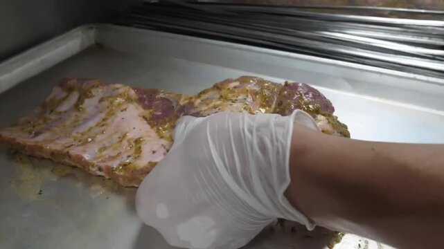 Close-up of a professional chef wearing a white glove seasoning a raw rack of pork ribs on a metal baking tray. The footage shows the preparation and marinating process of fresh meat in a commercial k