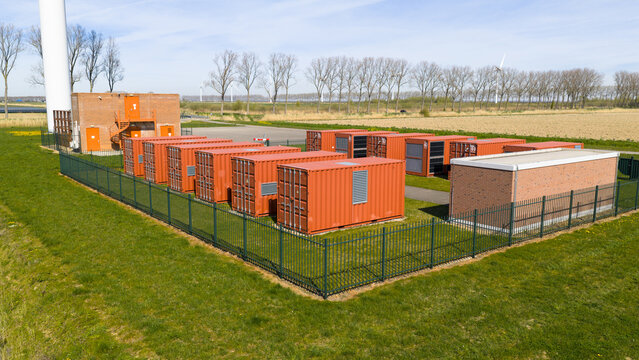 Aerial view of a battery energy storage system with orange containers and a brick utility building near a wind turbine in Middelharnis, Zuid-Holland, Netherlands.