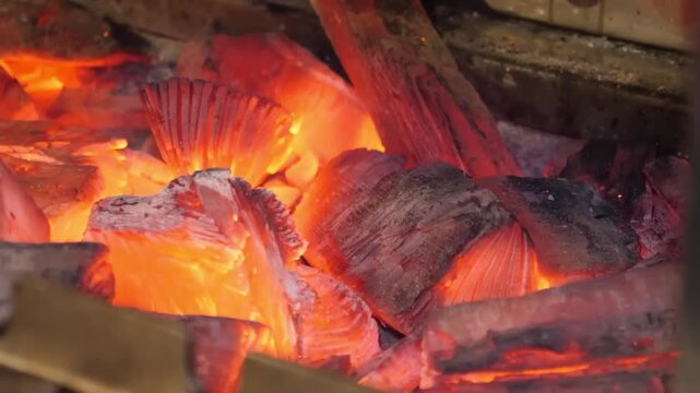 A macro shot captures the intense heat and vibrant orange and red glow of smoldering charcoal embers. The burning wood coals flicker with small sparks, providing a warm, fiery background texture.