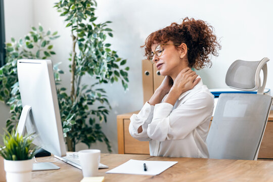 Tired mature woman with neck pain looking uncomfortable while working on a computer in the office