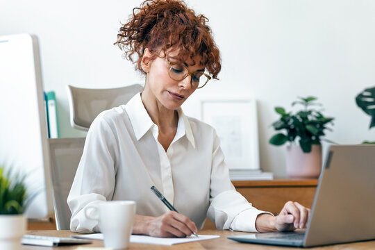 Concentrated mature business woman working with laptop while taking notes in the office