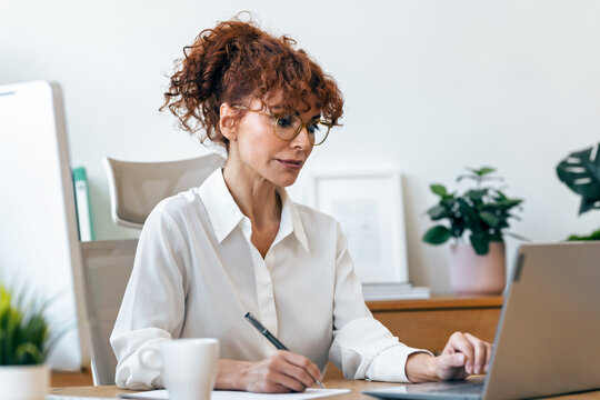 Concentrated mature business woman working with laptop while taking notes in the office