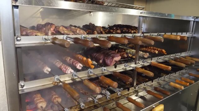 Close-up footage of various cuts of meat, including picanha steak, sausages, and skewers, rotating on a traditional rotisserie grill (churrasqueira) at a Brazilian steakhouse.