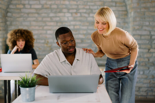 Diverse coworkers collaborating on laptop while discussing work tasks in contemporary office environment