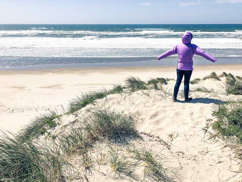 Rear view of a Woman in a puffer coat standing in the sand dunes on a sandy beach with her arms outstretched on a windy day, Florence, Oregon, USA
