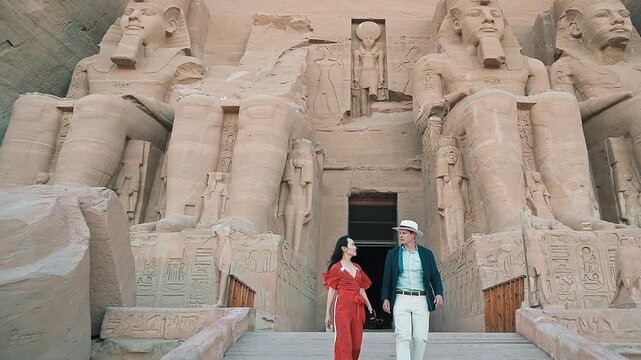american young man traveler wearing white hat with his wife with red dress exploring ancient Egyptian abu simble temple with background of ramsses ii statues. Egypt.