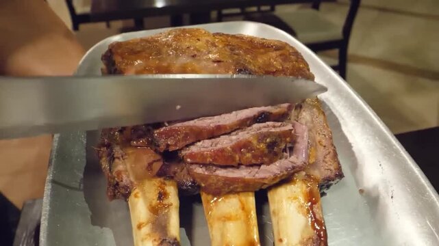 A close-up shot of a person's hand using a large knife to slice a piece of tender, slow-cooked beef short rib, revealing the succulent interior. The meat is served on a metal tray, highlighting the ri