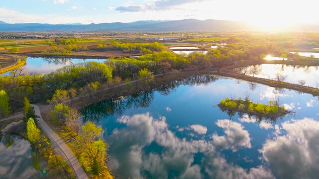 Aerial drone flies forward over glassy Colorado lakes reflecting clouds and spring foliage at golden sunset with Rocky Mountain backdrop and walking trail