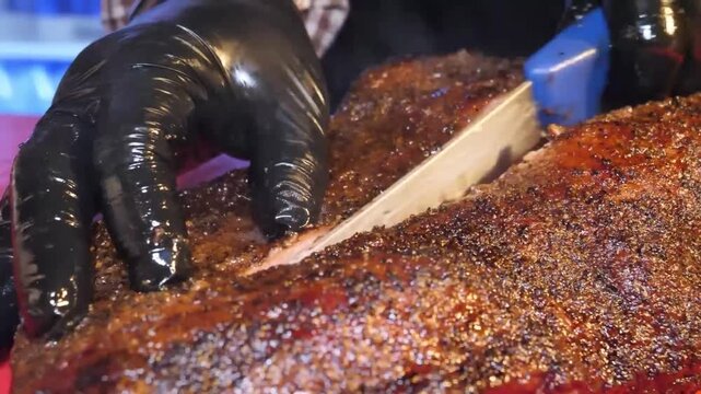 Close-up shot of a chef's gloved hands using a sharp, serrated knife to slice into a large, tender, and juicy smoked beef brisket with a dark, seasoned crust (bark). The meat is cooked low and slow fo