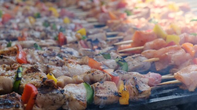 Cook at barbecue turns hot grilling kebab from lean meat pieces and vegetables on wooden eco skewers. Many barbecue at gastronomic festival. Aroma of roasted meat.