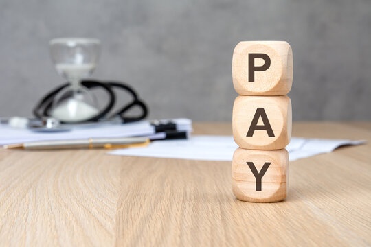 PAY word on vertically stacked wooden blocks. Blurred medical background with stethoscope and hourglass. Healthcare cost concept.