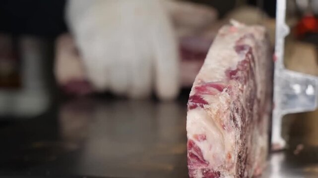 A close-up shot of a professional butcher wearing protective gloves using an electric industrial band saw to precisely cut through a large section of raw beef ribs and bone. The footage showcases the 