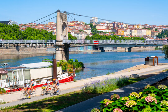 Motion blurred cyclists past along the sunlit banks of the Sa&ocirc;ne River, with the elegant suspension cables of the Passerelle Saint-Georges stretching toward the historic, vibrant buildings of Lyon's V