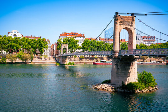 The Passerelle du Coll&egrave;ge suspension bridge spans the Rh&ocirc;ne River in Lyon, connecting stone piers to tree-lined quays. Its historic cables stretch across the blue water toward the vibrant cityscape.