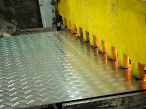 Heavy duty guillotine shears with yellow protective guard press down on silver diamond plate steel during precise industrial manufacturing cutting process in professional metalwork factory workshop.