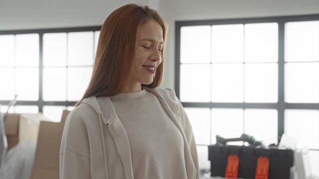 Woman carrying packages, smiling and looking sideways in building with cardboard boxes and a tool case visible; optimism.