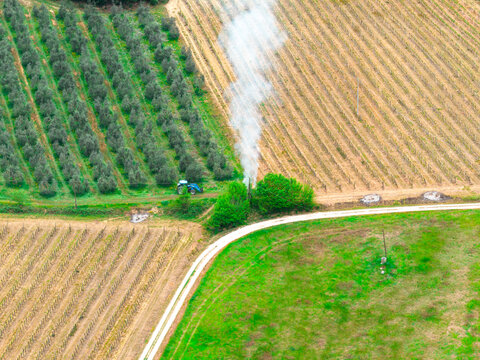 Aerial view of a blue tractor working near a plume of white smoke between olive groves and vineyards in Ficulle, Umbria, Italy.