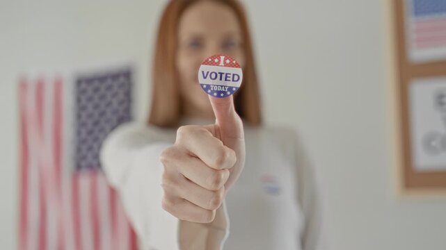 Woman redhead showing a voted sticker on her thumb, thumb extended toward camera with us flag and polling signage in a building; pride civic duty.