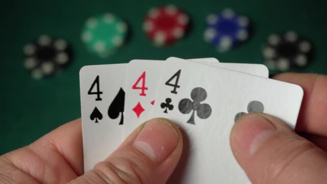 Close-up of hands gripping playing cards displaying three of a kind or set, with poker chips in green, red, and blue blurred in the background, emphasizing the card game setting