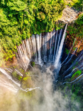 Aerial view of Tumpak Sewu Waterfall with multiple streams cascading down rocky cliffs into a misty canyon surrounded by lush jungle in Kecamatan Pronojiwo, Jawa Timur, Indonesia.